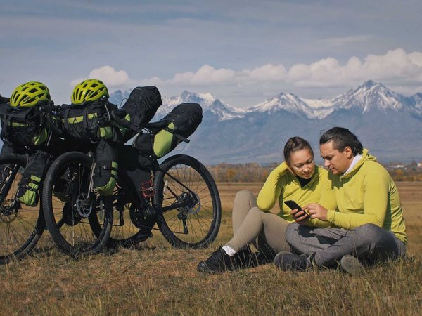 The man and woman travel on mixed terrain cycle touring with bikepacking. The two people journey with bicycle bags. Mountain snow capped.