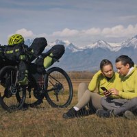 The man and woman travel on mixed terrain cycle touring with bikepacking. The two people journey with bicycle bags. Mountain snow capped.