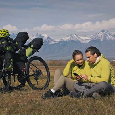 The man and woman travel on mixed terrain cycle touring with bikepacking. The two people journey with bicycle bags. Mountain snow capped.