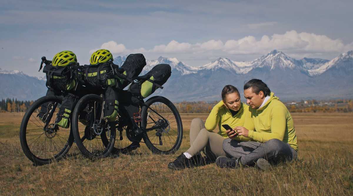 The man and woman travel on mixed terrain cycle touring with bikepacking. The two people journey with bicycle bags. Mountain snow capped.